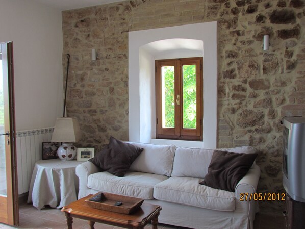 Un salón con pared de piedra, un sofá blanco, una mesita con lámpara y una ventana con contraventanas de madera.