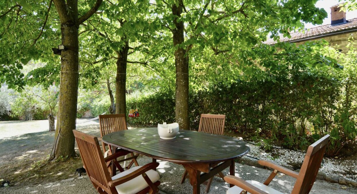 An outdoor dining area with a round table and chairs, surrounded by trees and greenery.