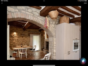 Una cocina rústica con paredes de piedra, vigas de madera, un cesto colgante y un comedor con mesa y sillas.