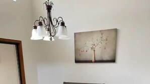 Uma sala de jantar com uma mesa quadrada, quatro cadeiras, um candelabro e um quadro na parede.