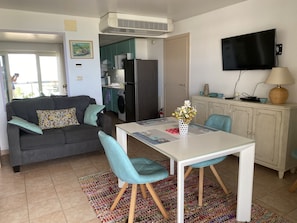 Un salon moderne avec un canapé gris, des fauteuils turquoise, une table blanche et un coin cuisine comprenant un réfrigérateur, une cuisinière et un téléviseur.