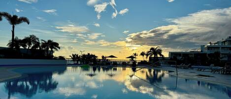 Una piscina con agua azul cristalina que refleja el cielo, rodeada de tumbonas y palmeras, con vistas a la puesta de sol.