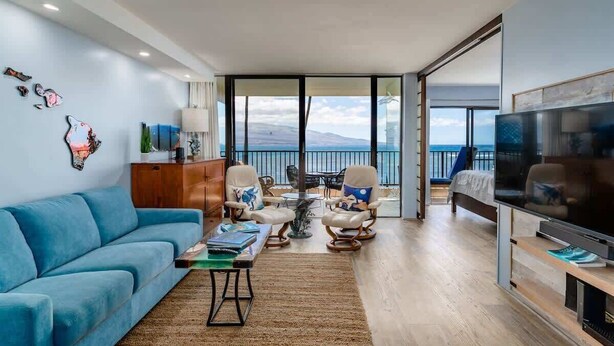 A modern living room with a blue sofa, two armchairs, a coffee table, and a TV. There is a balcony with a view of the sea and mountains.