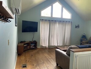 Un salon avec un téléviseur, un plancher en bois, une fenêtre avec des rideaux, un canapé et une petite étagère.