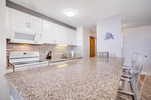 A kitchen with white cabinets, a granite countertop, a microwave, a stove, and a dining area with a glass table and chairs.