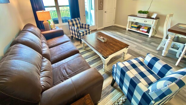 A living room with a brown leather sofa, blue and white checkered chairs, a wooden coffee table, and a TV mounted on the wall. - NEW! SPACIOUS condo—STUNNING views & CLOSE to Strip, SDC & LAKE! Sleeps 12!! (Branson)