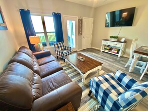 A living room with a brown leather sofa, blue and white checkered chairs, a wooden coffee table, and a TV mounted on the wall.