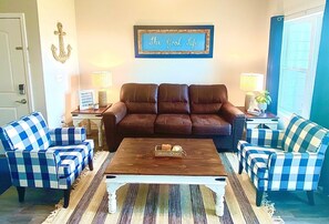A living room with a brown leather sofa, two blue and white checkered armchairs, a wooden coffee table, and a framed sign on the wall.