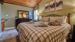 A bedroom with a wooden ceiling, a large bed with a checkered bedspread, a dresser, a mirror, and two wall-mounted paintings. - DOG FRIENDLY! Less than a mile from Dollywood and a min drive to the Parkway (Pigeon Forge)