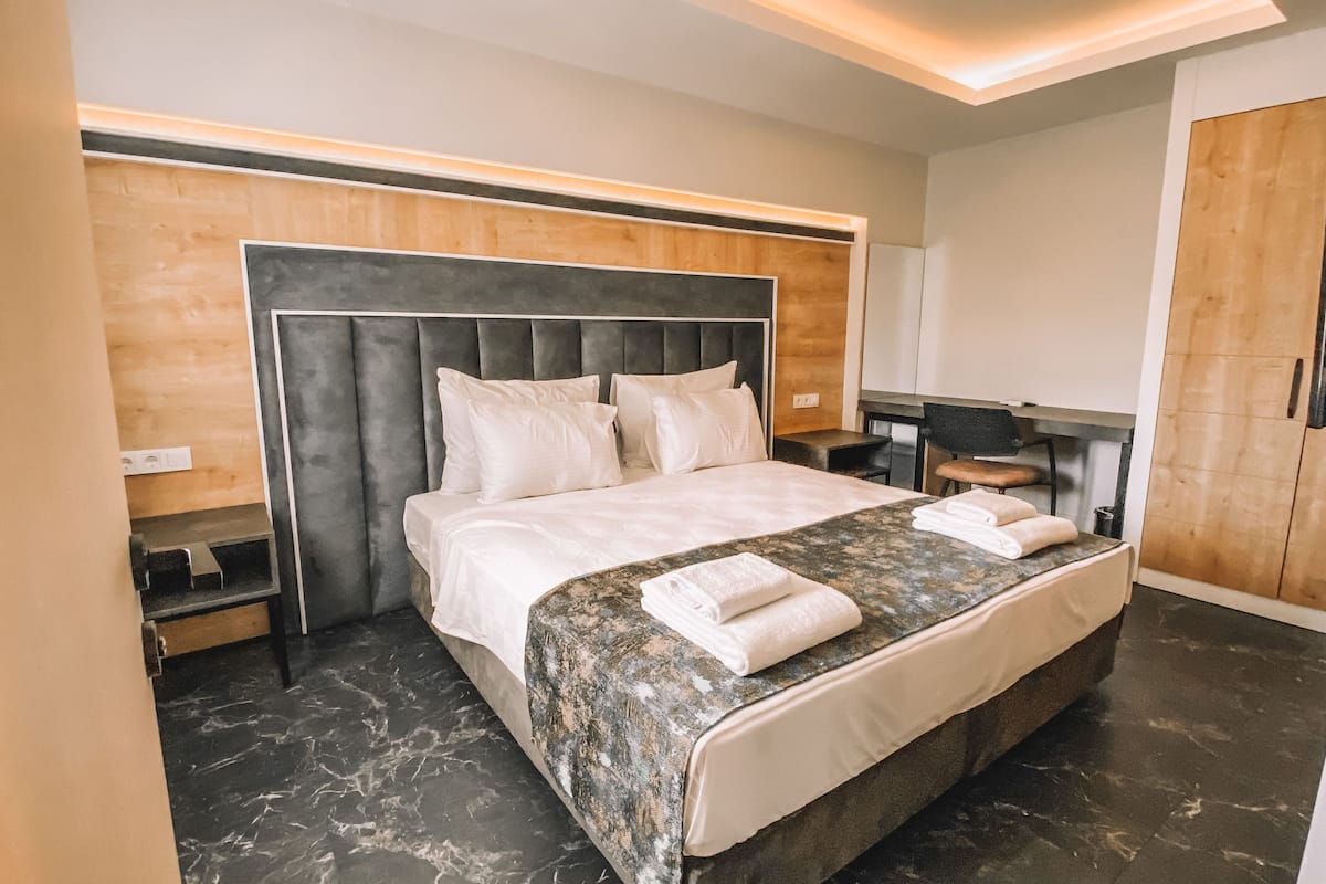 A modern hotel room with a large bed, a wooden headboard, a marble floor, and a small desk with a chair.