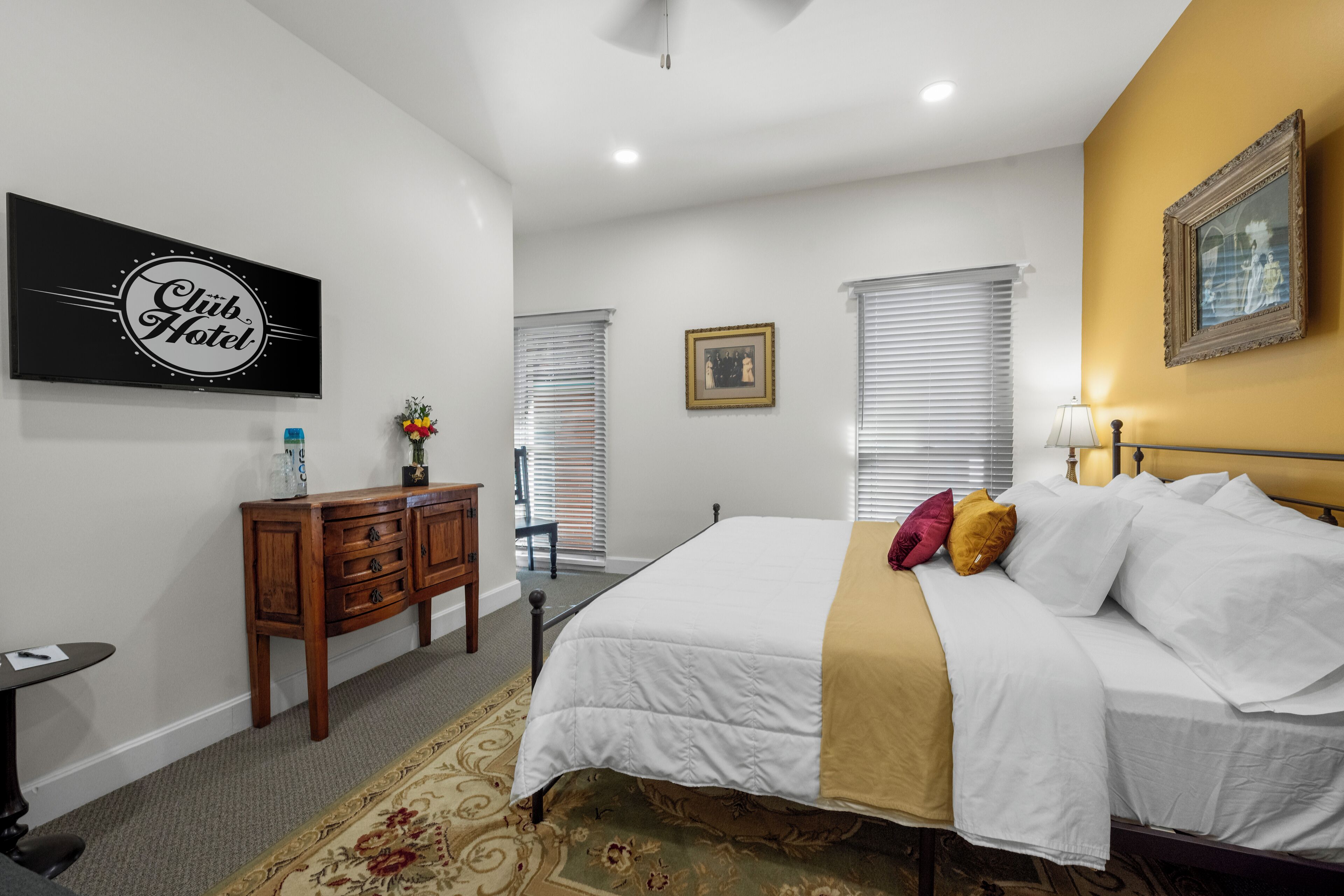 Une chambre d’hôtel comprenant un lit, une table de chevet, un bureau, une chaise, une fenêtre avec des stores, un tableau encadré au mur et une pancarte sur le mur.