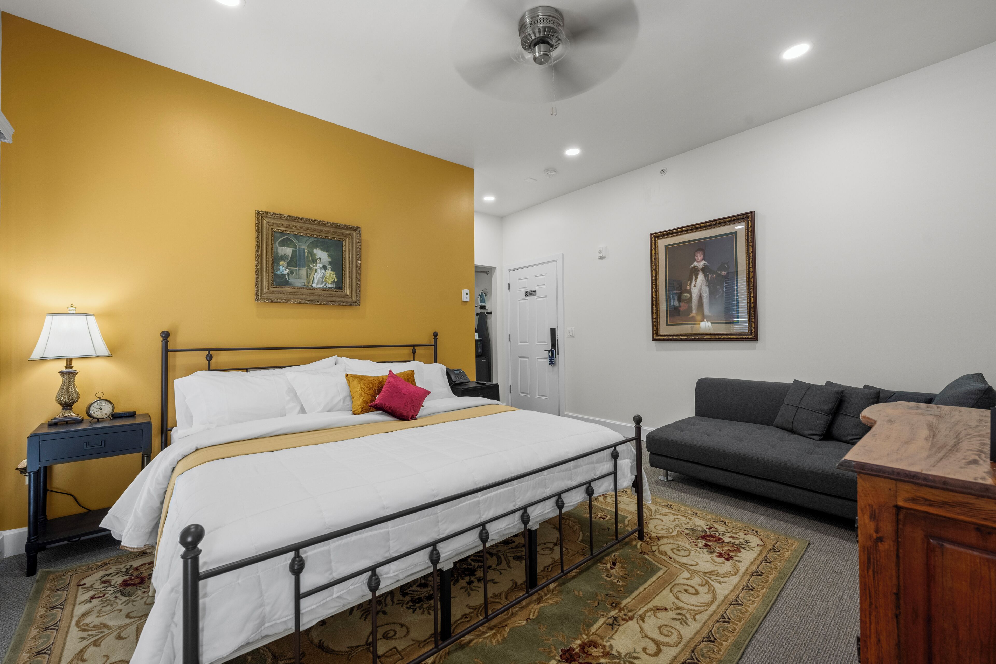 A bedroom with a bed, a nightstand, a lamp, a gray sofa, and two framed pictures on the walls.