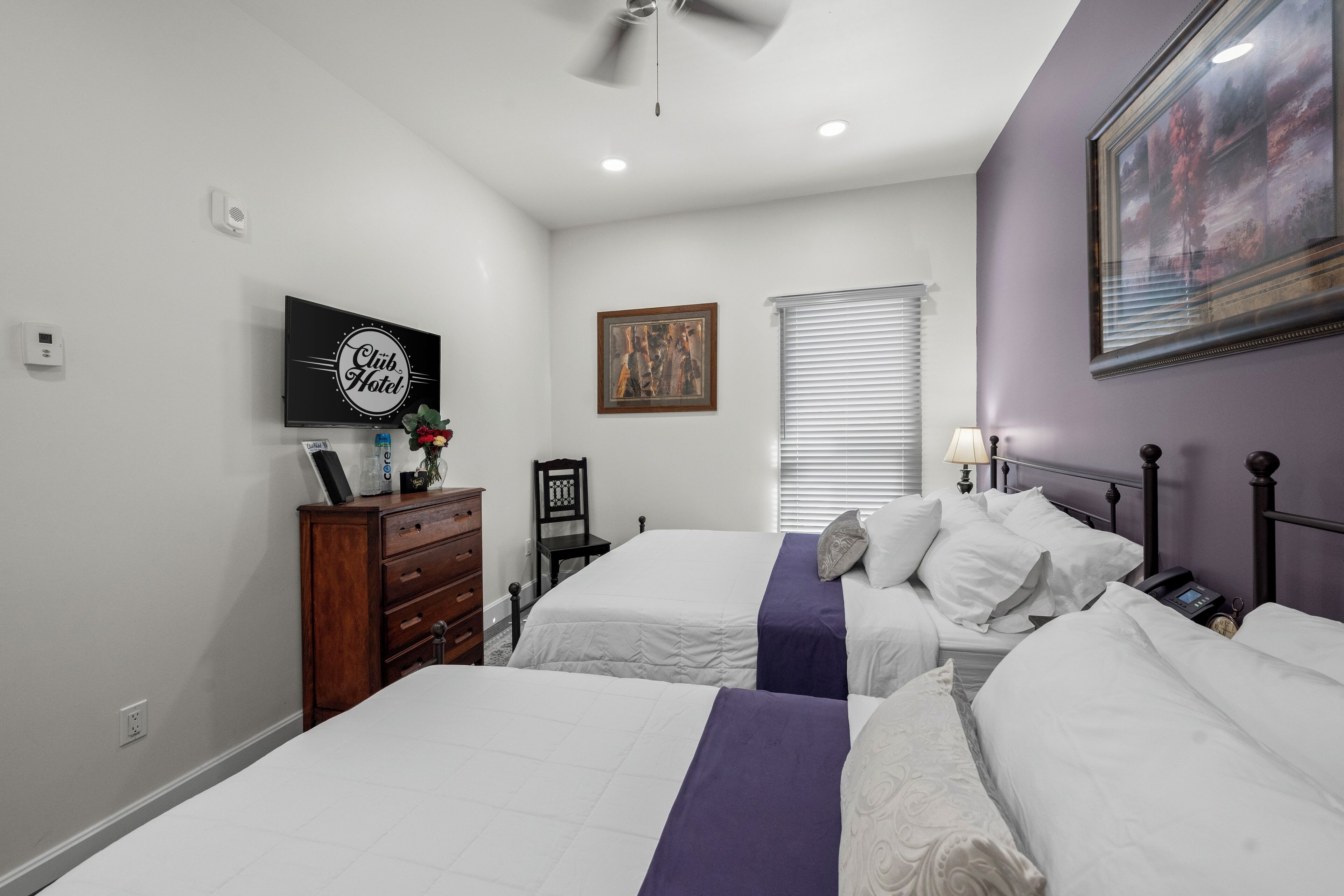 A hotel room with two beds, a dresser, a television, a painting, and a window with blinds.