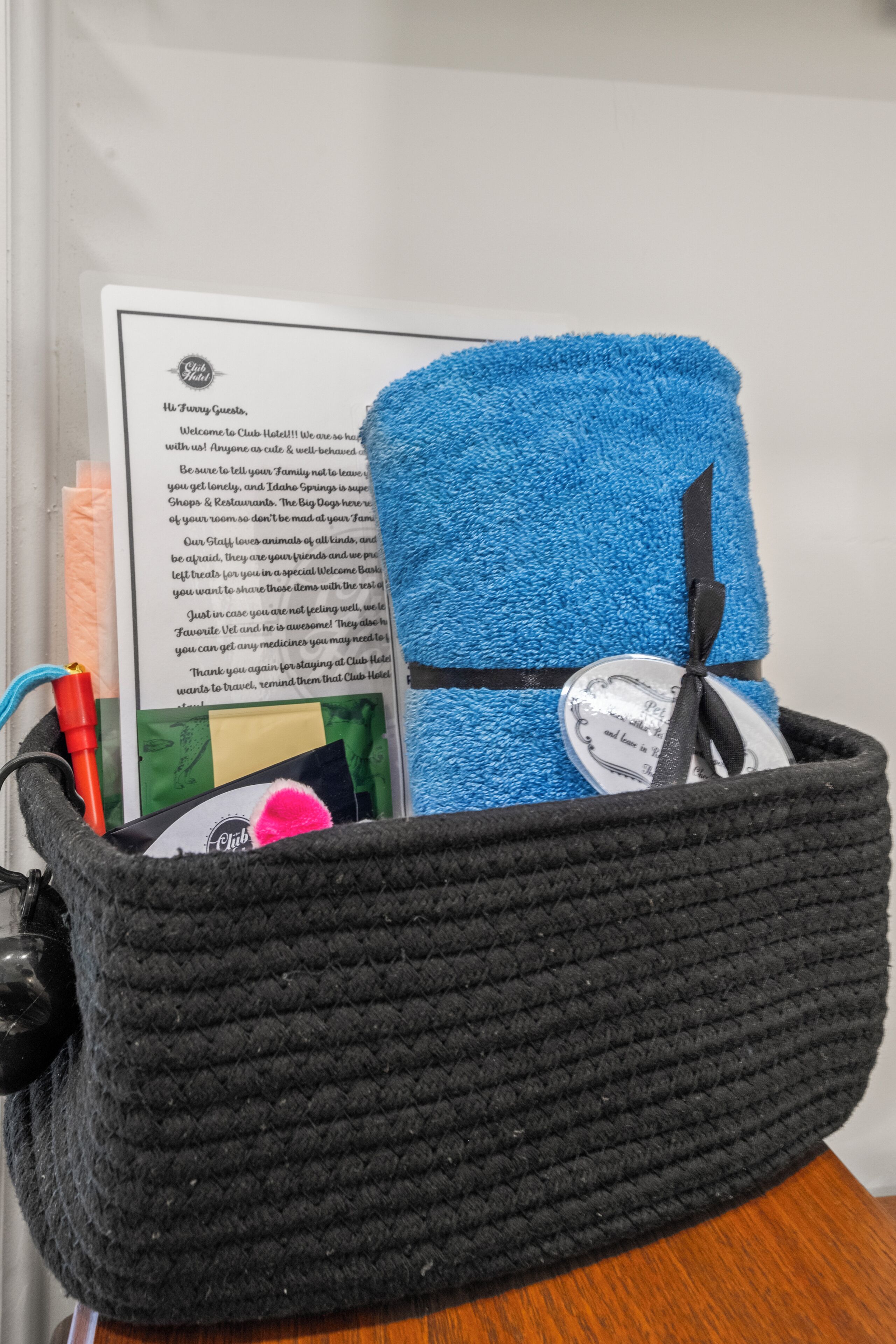 A black woven basket containing a blue towel, a black ribbon, a pen, and a printed welcome card.