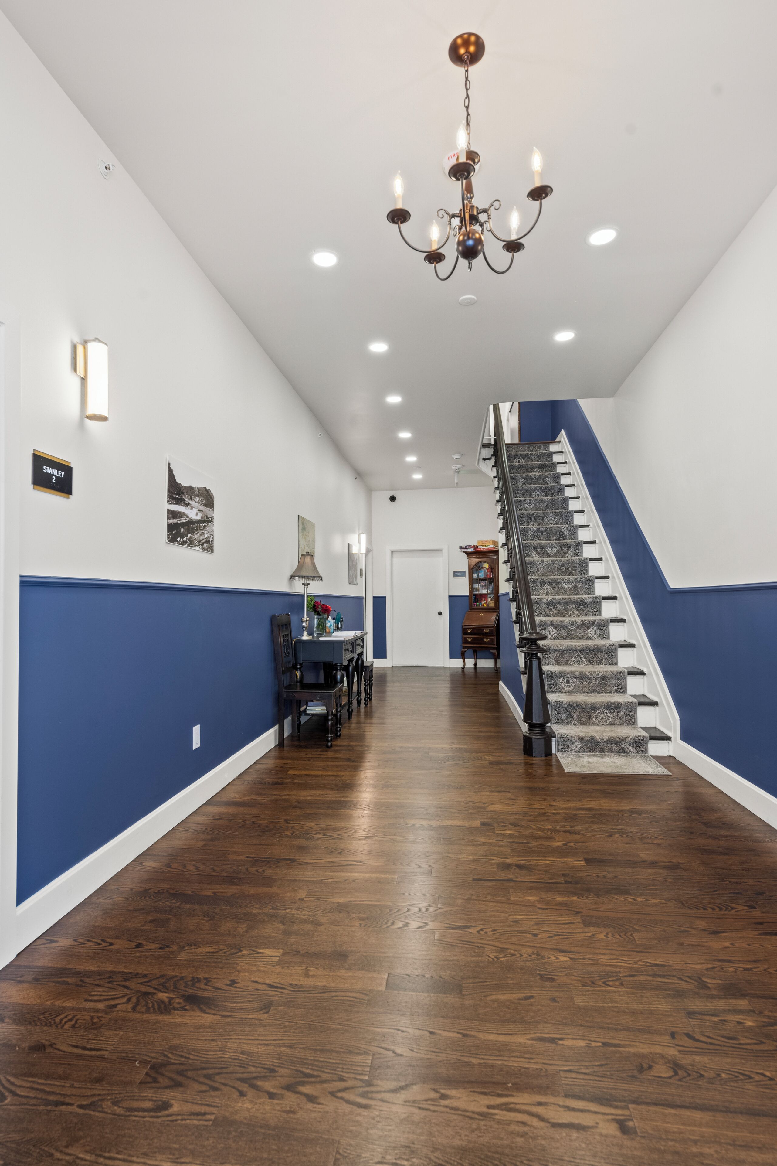 A hallway with a staircase, wooden flooring, a chandelier, and wall-mounted lights.