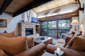 A cozy living room with a stone fireplace, wooden ceiling beams, and a large sliding glass door leading to an outdoor deck.