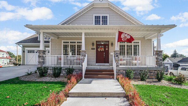 A two-story house with a front porch, stone pillars, and a red flag.