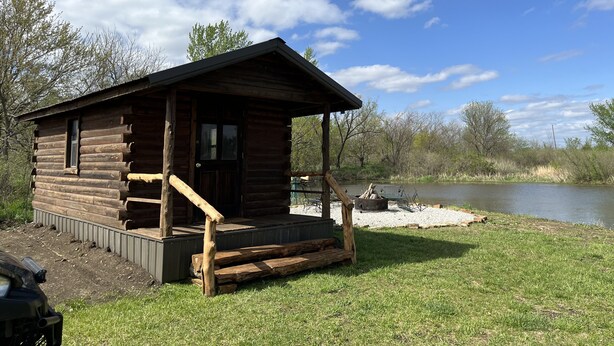 Un chalet rustique en rondins de bois, doté d’un porche et d’un foyer extérieur, situé au bord d’un lac.