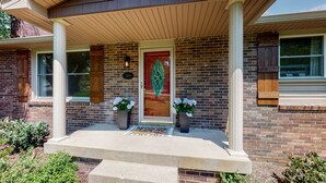 Une maison en briques avec un porche à l’avant, deux plantes en pot et une porte décorative.