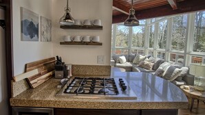 Une cuisine dotée d’un plan de travail en granit, d’une cuisinière à gaz et d’un coin repas offrant une vue sur les arbres à travers de grandes fenêtres.