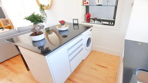 Et kompakt køkken med vaskemaskine, kaffemaskine og et lille bord med en potteplante.