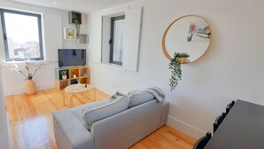 En moderne stue med en grå sofa, et lille rundt bord, et fjernsyn og et spejl monteret på væggen.