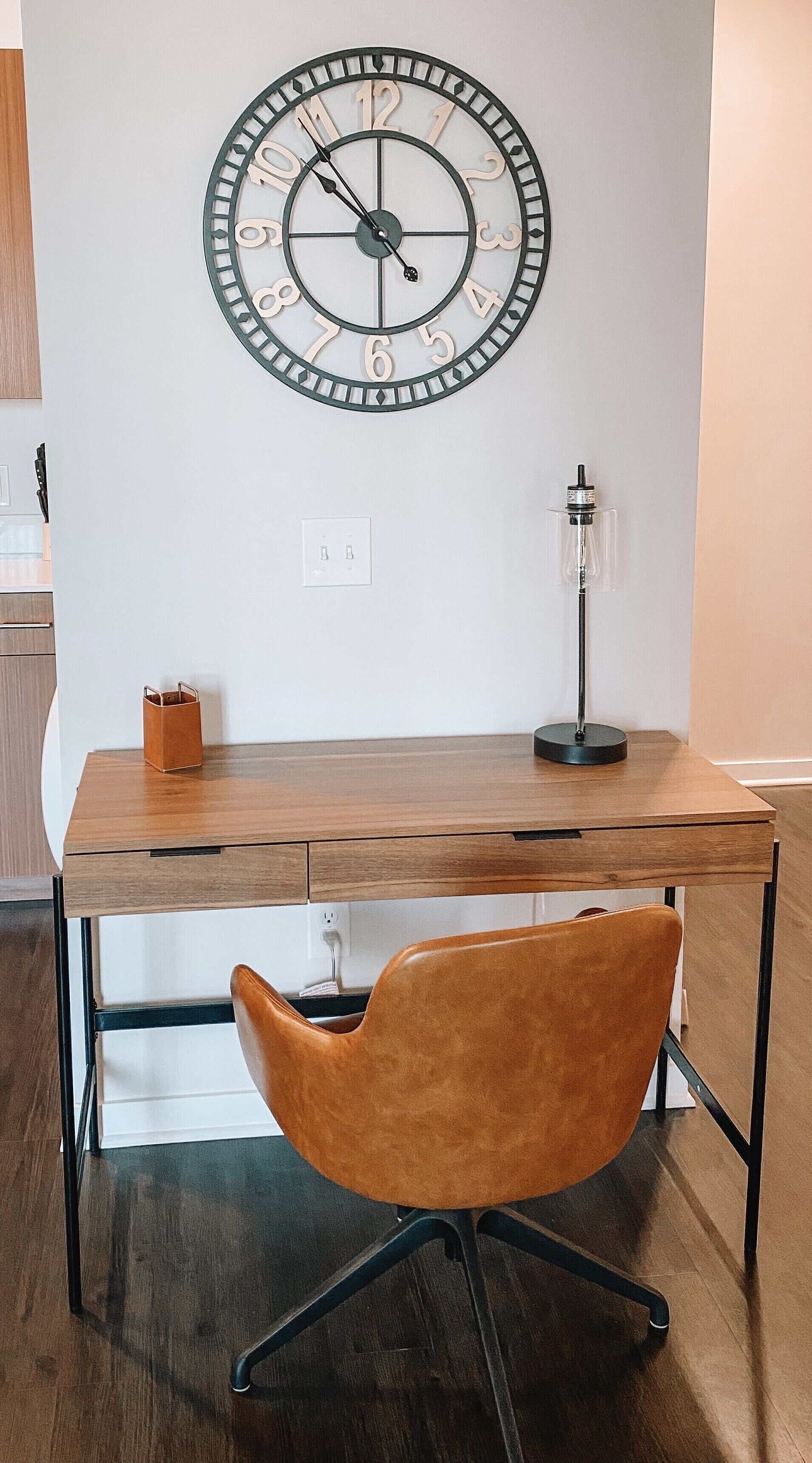 Un bureau en bois, avec une horloge, une lampe et une chaise, dans une pièce aux murs blancs et au plancher en bois.