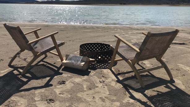 Two wicker chairs and a basket on a sandy beach with a body of water and mountains in the background. - Firepit-Shade-Chairs-Dark Skies (Levan)