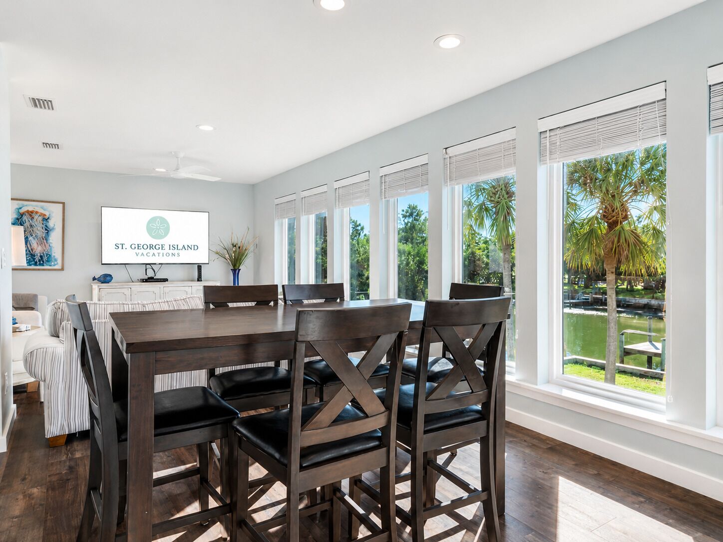 Un espace repas moderne avec une grande table et des chaises, un téléviseur diffusant des images de « St. George Island Vacations », et de grandes fenêtres offrant une vue sur la verdure.