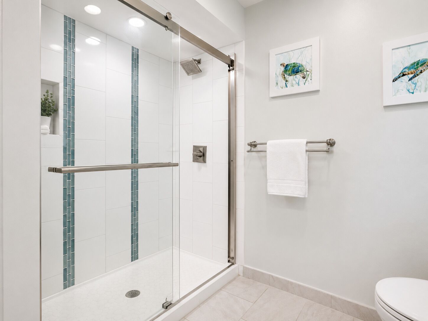 A modern bathroom with a glass-enclosed shower, white tiles, a towel rack, and two framed artworks on the wall.