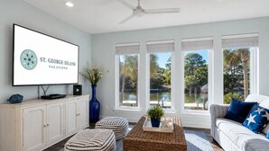Un salon avec un téléviseur affichant « VACANCES À ST. GEORGE ISLAND », un meuble blanc, un vase bleu, une table basse en osier et un canapé rayé agrémenté de coussins en forme d’étoile.