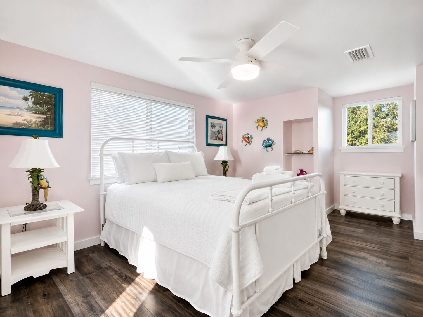 Un dormitorio con cama, mesitas de noche, ventilador de techo, ventana con persianas y decoración en las paredes.