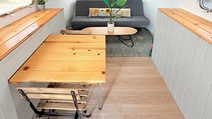 Een ruimtelijk ingericht camperinterieur met een houten tafel, een bank met kussens, een kleine plant en een raam.