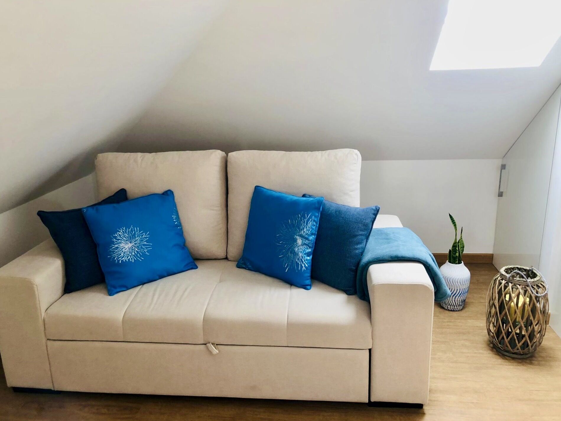 A beige sofa with blue pillows, a small potted plant, and a woven basket in a room with a sloped ceiling and wooden flooring.