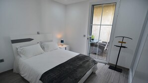 A modern hotel room with a large bed, a bedside table with a lamp, a sliding glass door leading to an outdoor area with a table and chairs. - Casas do Baleeiro - Whaler’s Houses (Machico)