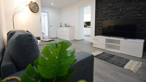 A modern living room with a flat-screen TV, a grey sofa, a white entertainment unit, and a stone accent wall. - Casas do Baleeiro - Whaler’s Houses (Machico)
