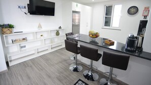 A modern kitchen with a flat-screen TV, white cabinetry, and a stainless steel refrigerator. There are three bar stools and a fruit bowl on the counter. - Casas do Baleeiro - Whaler’s Houses (Machico)