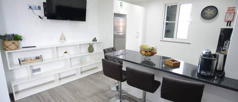 A modern kitchen with a flat-screen TV, white cabinetry, and a stainless steel refrigerator. There are three bar stools and a fruit bowl on the counter.