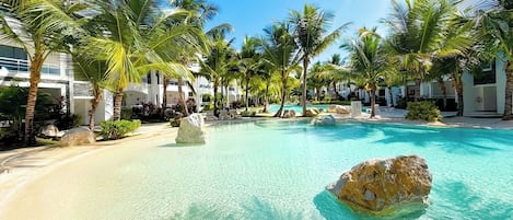 A resort pool area with clear turquoise water, palm trees, and large rocks.