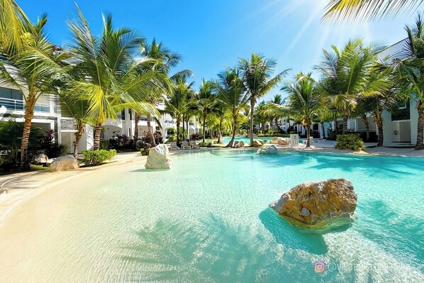 A resort pool area with clear turquoise water, palm trees, and large rocks.