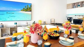 Comedor con vista a una playa tropical, una mesa de madera decorada con frutas, bebidas y un florero.