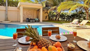 A poolside dining area with a table set for two, featuring tropical fruits, drinks, and a cozy seating area with a pool in the background.