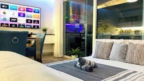 Een hotelkamer met een grote televisie waarop diverse streamingdiensten worden getoond, een bed met een gestreept kussen, een bureau met een stoel, en een uitzicht op de stadshemel door het raam.