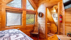 Een gezellig houten vakantiehuisje van binnen, met een groot raam, een houten stoel, een bed met sprei en een open haard.