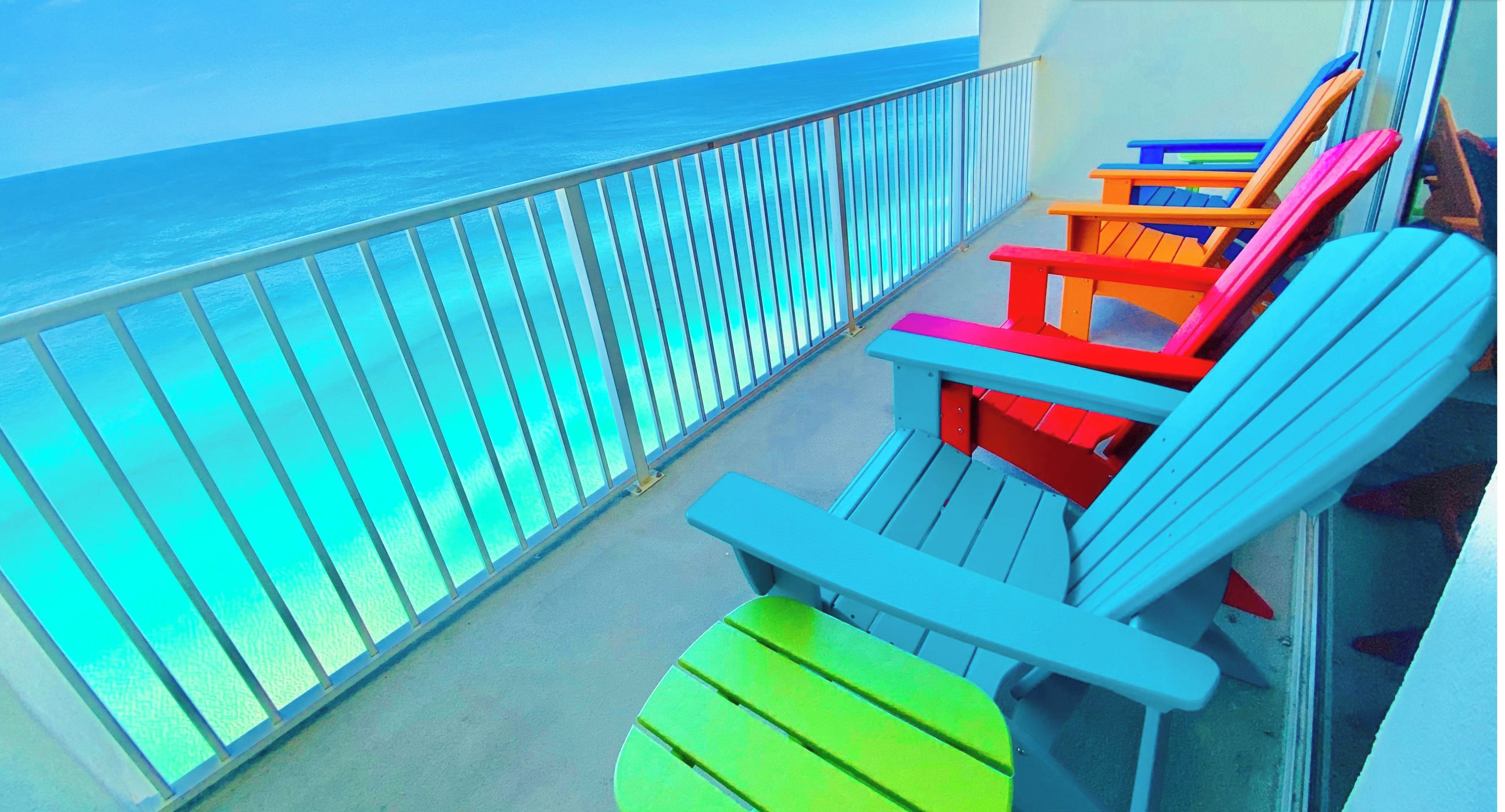 Un balcon offrant une vue sur l’océan, agrémenté de chaises Adirondack aux couleurs vives et d’une rambarde en verre.