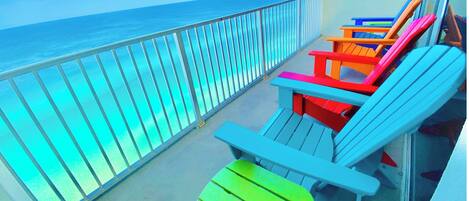 A balcony with a view of the ocean, featuring colorful Adirondack chairs and a glass railing.