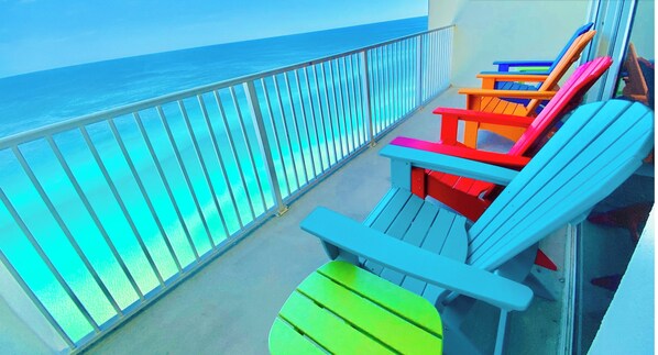A balcony with a view of the ocean, featuring colorful Adirondack chairs and a glass railing.