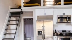 Una cocina moderna con electrodomésticos de acero inoxidable, un comedor con mesa de madera y sillas metálicas, y una escalera que sube a un nivel superior.