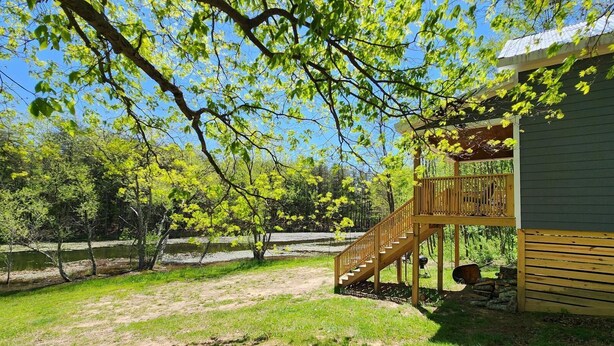 Una terraza de madera con una escalera que lleva a una zona cubierta, rodeada de árboles y un césped.