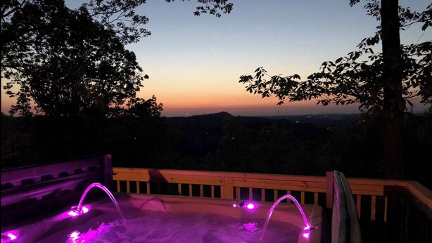 A hot tub with purple lighting, surrounded by a wooden deck, overlooking a scenic landscape during twilight. - MOUNTAIN TOP Lodge with Gorgeous Panoramic View, Hot Tub, Pool Table & Fireplace (Sevierville)